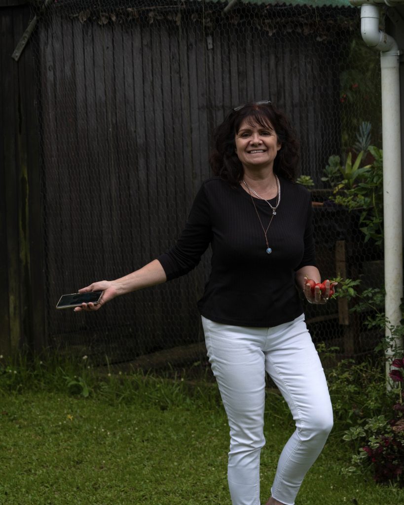 A smiling woman standing in a colourful cottage garden surrounded by flowers, plant pots and greenery on a sunny morning.
