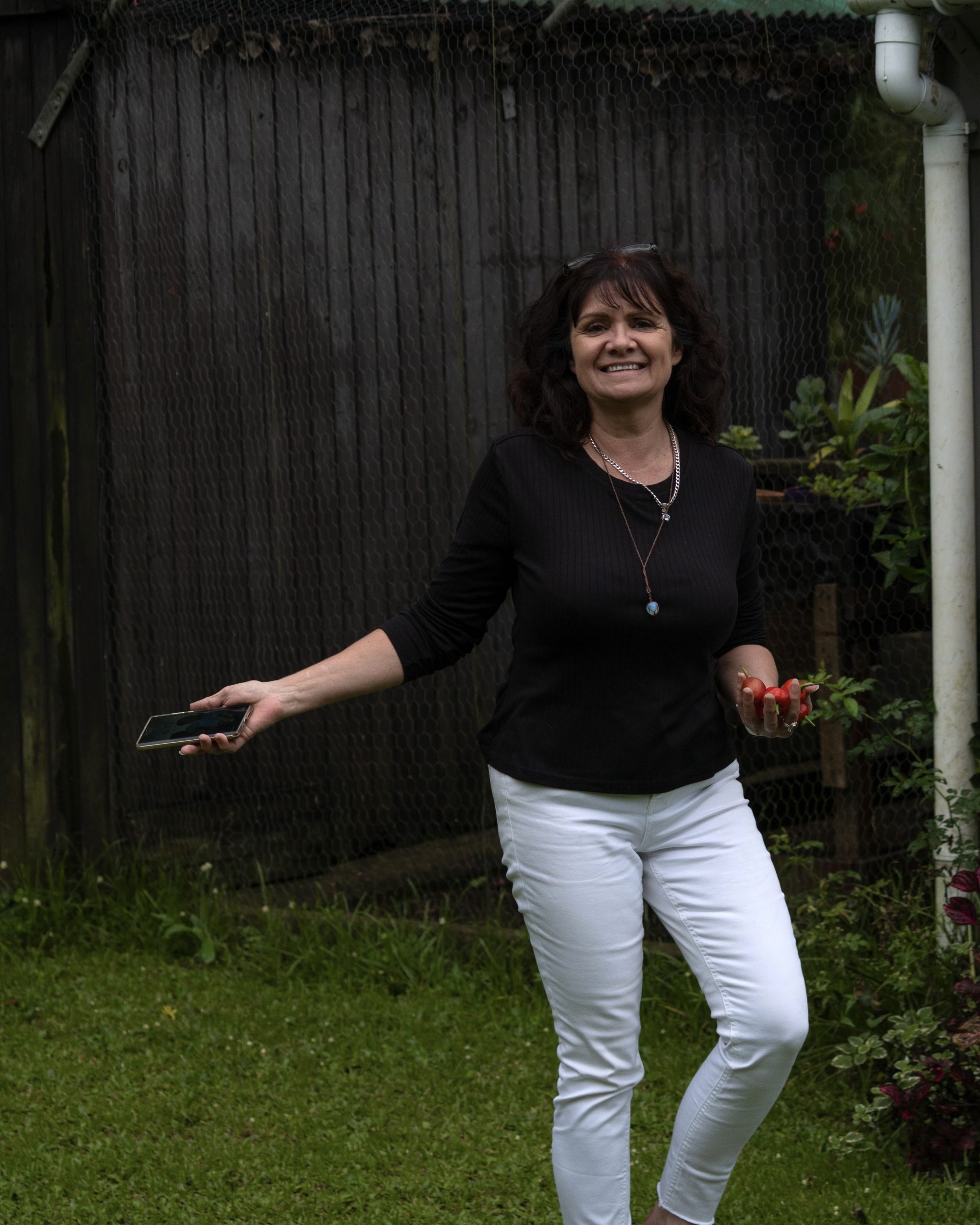 A smiling woman standing in a colourful cottage garden surrounded by flowers, plant pots and greenery on a sunny morning.